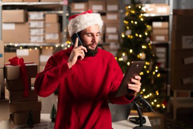 Lojistik, nakliye ve depo işçisi telefonda tabletle konuşuyor. Akıllı telefonu olan bir adam Noel 'de depoda nakliye ve uluslararası dağıtım işi yapıyor. Boşluğu kopyala