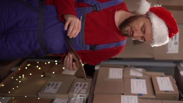 サンタ帽子をかぶっている男性労働者は小売倉庫でデジタルタブレットで在庫と在庫をチェックします 配送センターでクリスマスに取り組んでいます バーティカルビデオ — ストック動画