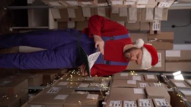 Lojistik ithalat konsepti. Depodaki paket kutusunu kontrol eden genç bir işçi. Depoda çalışan beyaz bir adam pano tutuyor. Dikey video
