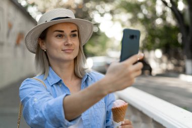 Sokakta yürüyen genç sarışın kadın portresi cep telefonuyla selfie çeker dondurma yer. Boşluğu kopyala