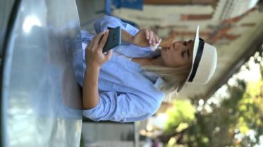 Dondurmalı şapkalı genç bir kadın arkadaşlarıyla iletişim kurmak için telefon kullanıyor. Kadın, giyinmiş gömleğiyle sokak kafeteryasında duruyor. Sosyal medya bağımlılığı kavramı. Dikey video