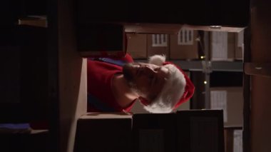 Noel 'de endüstriyel fabrika deposundaki raflarda pano kontrol ürünleri ya da paket ürünleri kullanan bir erkek çalışan. Noel Baba şapkalı envanter müdürü. Dikey video