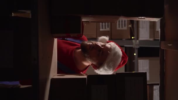男性员工仓库工作人员在工厂仓库使用剪贴板检查产品或货架上的包裹货物 圣诞老人帽的库存经理 垂直录像 — 图库视频影像