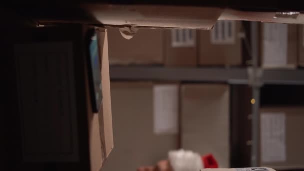サンタハットの倉庫労働者は 箱付きの棚に電話を探し 郵便局の同僚を呼び出しました バーティカルビデオ — ストック動画