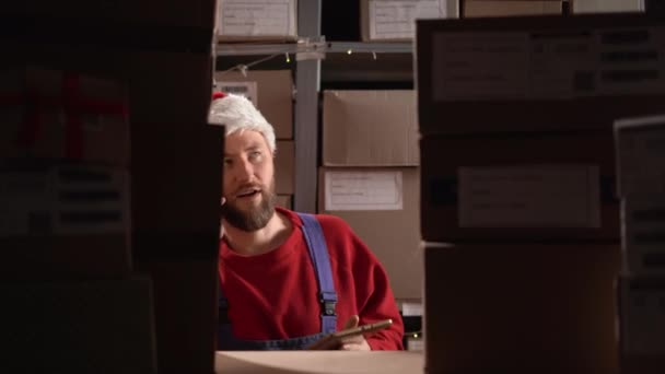 サンタの帽子をかぶっているハンサムな倉庫労働者は 在庫を確認するためにデジタルタブレットを使用し 彼は段ボール箱が付いている棚の近くの携帯電話で呼び出しています コピースペース — ストック動画