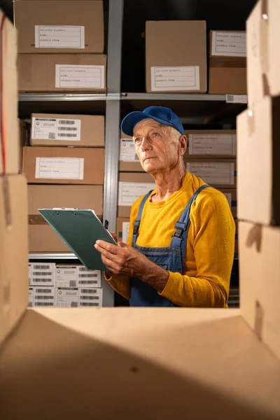 Elderly male inventory manager checks stock, writing notes in the clipboard. Senior man working in a warehouse storeroom with shelves full of cardboard boxes. Copy space