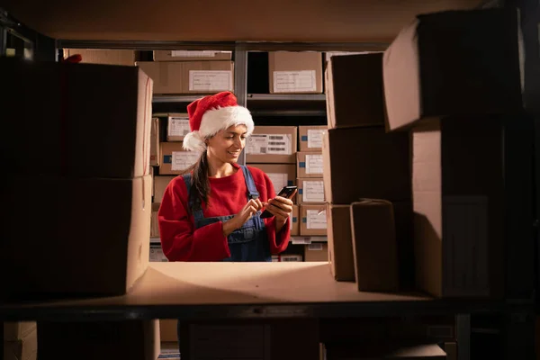 仓库工人用手机检查存货 员工在货架附近戴着圣诞帽 纸板箱 在仓库工作 复制空间 — 图库照片