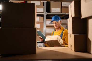 Yaşlı teslimatçı ya da depo çalışanı. Paket kutusu ve mavi önlüklü, mal geçmişi üzerinde. Lojistik iş ve sevkiyat konsepti. Boşluğu kopyala