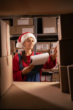 Bayan çalışan depo çalışanı. Fabrika deposundaki raflarda pano kontrol ürünleri ya da paket ürünleri kullanıyor. Noel Baba depoda çalışıyor.