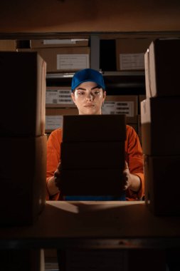 Depoda çalışan kadın, karton kutuları rafa koyar. Raflarda sevkiyata hazır ürünlerle dolu karton kutular ve paketler. Boşluğu kopyala