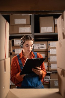 Rafın yanında duran kulaklık takan kadın profesyonel operatör depodaki paketleri sayıyor, malların envanterini çıkarıyor. Boşluğu kopyala