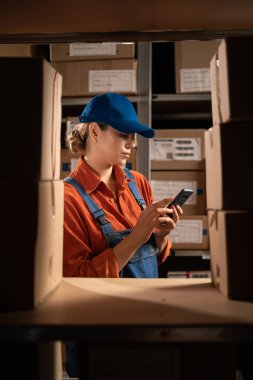 Mobil uygulama kullanan kadın bir depo çalışanı paket kutusunu kontrol ediyor. Girişimci elinde cep telefonu teknolojisi cihazıyla paketlerle rafların yanında dikilirken. Boşluğu kopyala