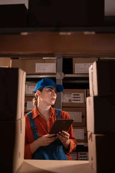 Manager checking inventory uses digital tablet. Young woman working in warehouse with rows of shelves full of parcels. Copy space