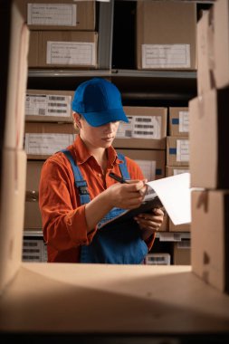 Endüstriyel fabrika deposunda çalışan kadın bir depo çalışanı pano kullanarak raflardaki eşyaları kontrol ediyor. Denetleme kalite kontrolü. Boşluğu kopyala