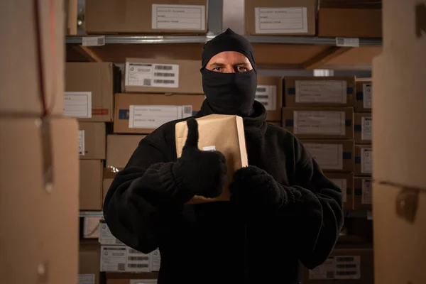 Adult male thief an in a ski mask picks up a box with a parcel in a warehouse at night looking at camera and showing class sign. Copy space