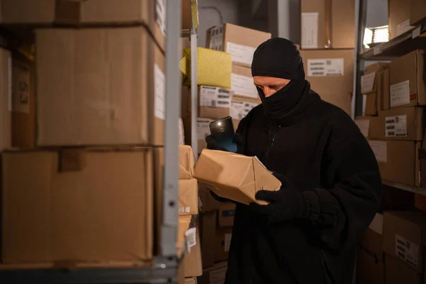 Male thief in black mask steals a parcel in a warehouse at night lightning with flashlight. Security problems in warehouses concept