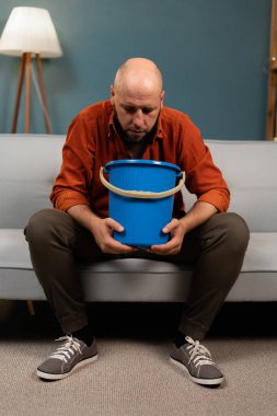 Kel adam midesi bulanıyor ve kusuyor. Elinde kovayla kanepede oturuyor. Boşluğu kopyala