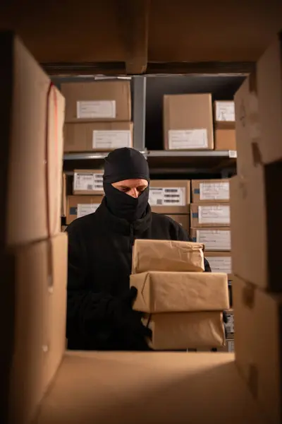 Thief in black balaclava steals with a parcel in a warehouse at the night. Concept of security problems in storehouses and stores. Copy space
