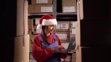Depoda dizüstü bilgisayarla çalışan Noel Baba şapkalı bir kadın işçi. İnsanlar, depo ve tatil konsepti
