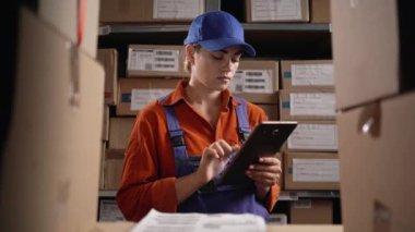 Depo çalışanı kutularla rafların yanında duran stokları kontrol etmek için dijital tablet kullanıyor. Boşluğu kopyala