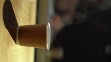 Bir bardağın yakın plan çekimi, içinde kahve olan bir bardak, buharlı ahşap bir masanın üzerinde duruyor ve bir kadın eli onu alıyor, bir kahve dükkanında sabah kahvesi kavramı. Dikey video