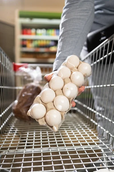 a stiff hand places a tray of eggs into a supermarket shopping cart. food purchasing concept.