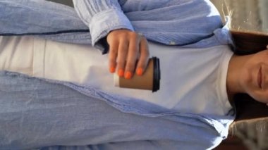 Şehir caddesinde yürürken gülümseyen ve kahve içen neşeli beyaz kadın, yakın plan. Dikey video