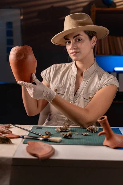 Female archaeologist working in the office at night holds an antique vase in her hands. Copy space