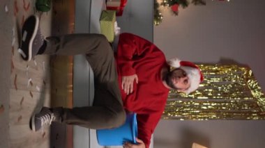 Kanepede Noel Baba şapkalı bir adam elinde bir kova tutuyor ve karnını tutuyor. Kusuyor. Noel 'de veya yeni yıl kutlamalarında gıda zehirlenmesi konsepti. Uzayı kopyala dikey video