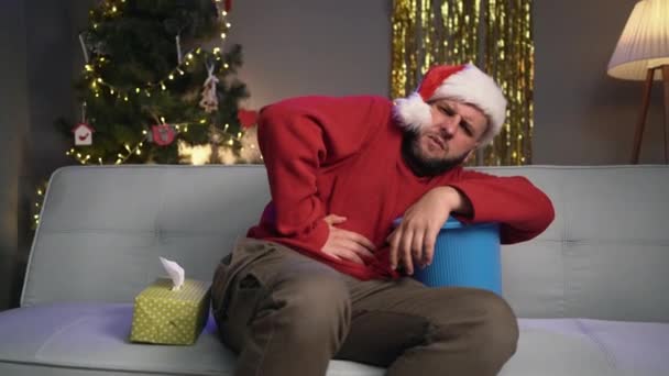 ある男性がサンタクロースの帽子をかぶったソファーに座り バケツを手に持って胃を抱えています クリスマスや新年のお祝いの間に食べ物中毒の概念 コピースペース — ストック動画