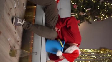 Kanepede Noel Baba şapkalı bir adam elinde bir kova tutuyor ve karnını tutuyor. Kusuyor. Noel 'de veya yeni yıl kutlamalarında gıda zehirlenmesi konsepti. Uzayı kopyala dikey video