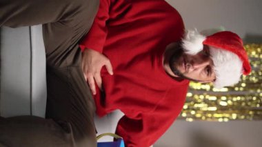 Kanepede Noel Baba şapkası takmış elinde kova tutan ve karnını tutan bir adam kusuyor. Noel 'de ya da yeni yılda gıda zehirlenmesi kavramı. Yakın plan portre. dikey video
