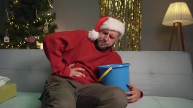Bir adam kanepeye oturmuş Noel Baba şapkası takıyor elinde kovayla karnını tutuyor. Noel 'de veya yeni yıl kutlamalarında gıda zehirlenmesi konsepti. Boşluğu kopyala.