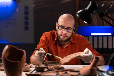 Arkeolog gece geç saatlerde ofiste çalı çırpıyla kil tabağı temizliyor. Boşluğu kopyala