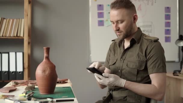 在办公室里研究古代文物的男性考古学家用智能手机坐在桌子旁边 复制空间 — 图库视频影像
