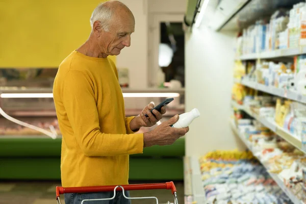Old man taking dairy products from shelf in the supermarket, holding bottle and smartphone, scanning barcode on product. Copy space