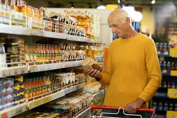 Elderly man in supermarket buying groceries food holding glass jar of salad walking along the aisle in grocery store. Copy space