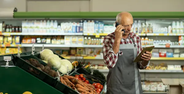 Elderly male sales assistant in a grocery using digital tablet and talking on cell phone while taking inventory. Banner. Copy space