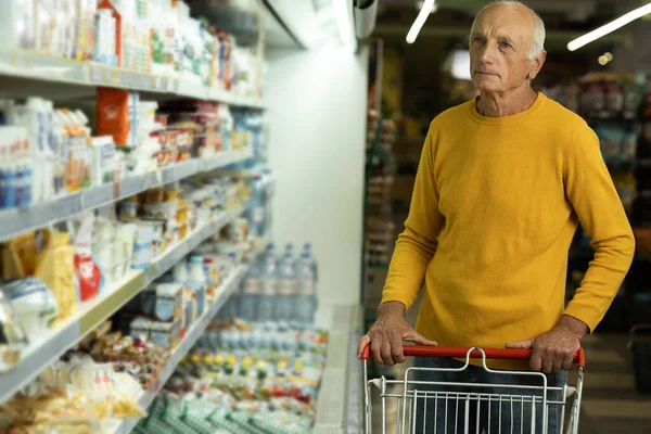 Old man standing with shopping trolley near the shelves in grocery store, buying dairy products in supermarket. Copy space