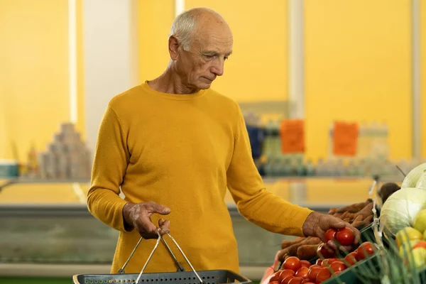 Old man choosing tomatoes from vegetable baskets in supermarket while buying eco groceries. Copy space