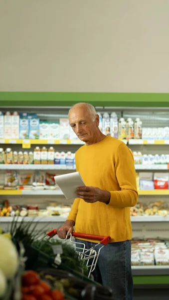 Elderly man at the supermarket shopping with a grocery list and pushing cart. Retail concept. Copy space