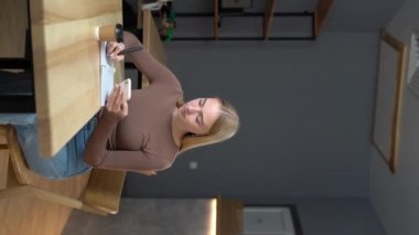 Kapalı kafedeki güzel sarışın kadın not defterine not tutarken cep telefonu kullanıyor. Dikey video