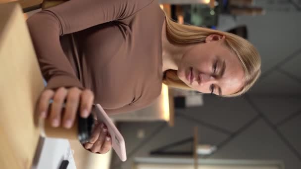 美しい若い女性がコーヒーの紙コップを持って カフェテリアに座っている間スマートフォンを見ています 携帯電話とコーヒーテイクアウトを飲んでいる少女 バーティカルビデオ — ストック動画