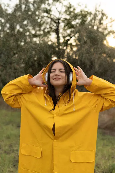 Young beautiful woman in a yellow raincoat enjoys music in the rain while walking in the park. Copy space