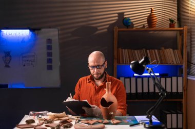 Gözlüklü sakallı bir arkeolog gece geç saatlere kadar ofiste çalışıp bilimsel işler yapan bir panoya not alıyor. Boşluğu kopyala