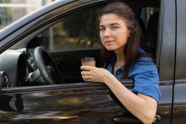 Mavi gömlekli güzel bir kadın kahve içiyor, kameraya bakıyor ve arabasında otururken gülümsüyor. Boşluğu kopyala