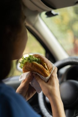 Hamburger yiyen ve arabasında oturan bir kadın. Arabada sağlıksız yemek. Boşluğu kopyala