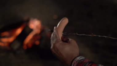 Adamın biri bir çubuğa sosis bağlayıp gece ateşte kızartıyor. Kamp ve kamp ateşi kavramı. Yakın plan.