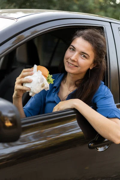 Woman driving car while eating hamburger enjoying takeaway food. Waiting and standing in traffic jam. Copy space