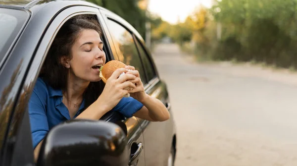 Beautiful woman eating a burger while driving, unhealthy lifestyle because eats of junk food. Banner. Copy space